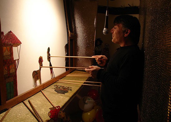 A person in a shadow play theater holding two shadow puppets close to a screen. 