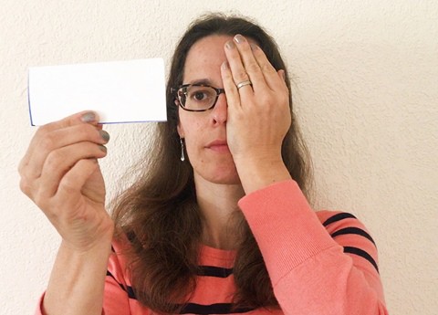A person covering her left eye with her left hand. In her right hand she holds a paper card close to her face at eye level.