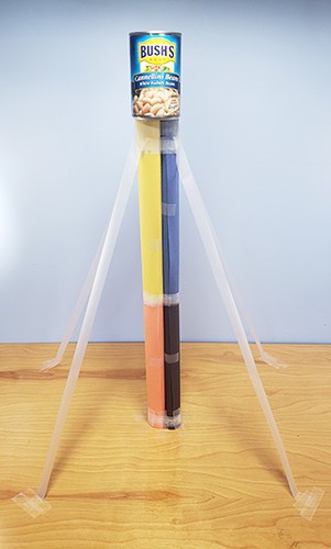 A paper tower made from vertical tubes with supporting guy wires made from tape, and a can of beans at the top.