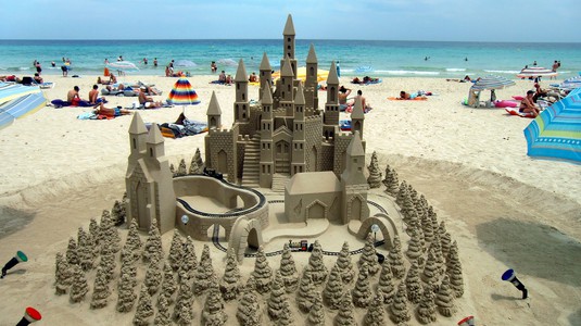 A large sandcastle with many spires, surrounded by a forest of sand trees and a train track. 