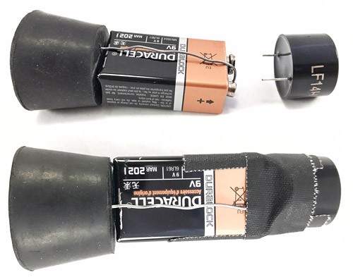 Leads of a buzzer are pressed onto the terminals of a 9 volt battery and taped in place