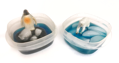 A model penguin and polar bear sit in plastic containers of blue ice water