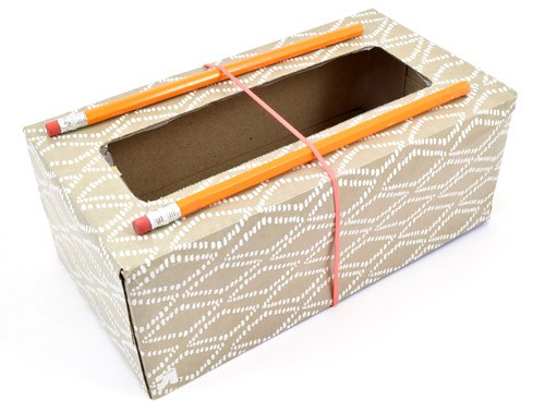 Two pencils lay parallel on opposite sides to the opening of an empty tissue box and are secured by a rubber band