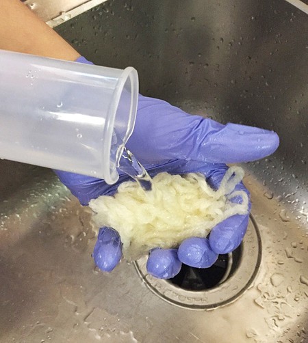   A gloved hand holding a piece of wool yarn in the sink. Water is poured over the wool from a measuring cylinder. 