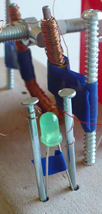 Two nails wrapped with magnet wires hold the leads of an LED in a wooden board