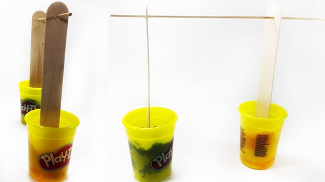 Front and side view of two small containers of modeling clay holding wide craft sticks  upright. A skewer pokes through the craft sticks, making a horizontal bar. 