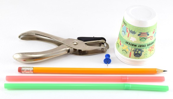 A hole puncher, stack of paper cups, thumb tack, pencil and two plastic straws