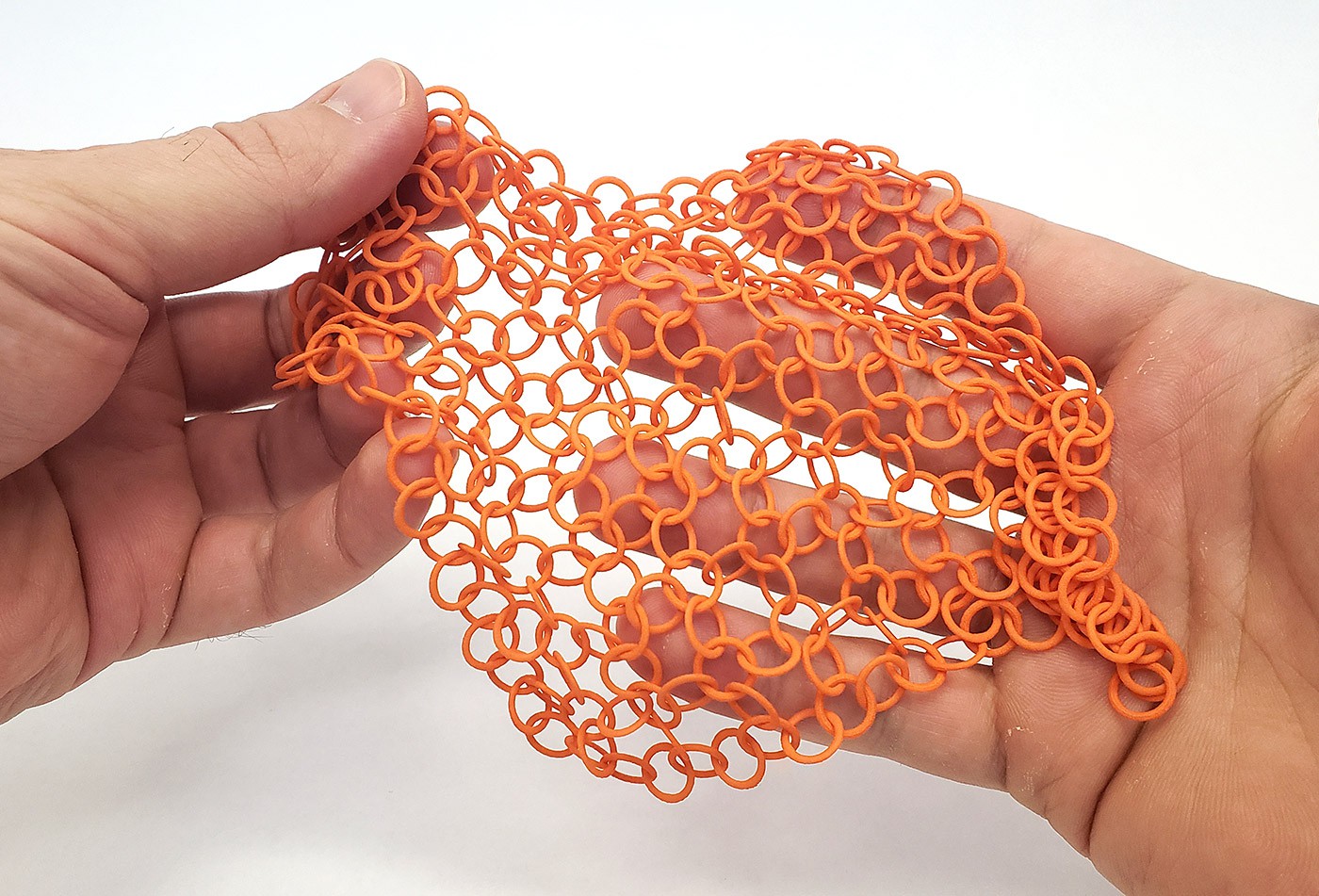 Two hands hold a piece of 3D printed fabric. The interlocking ring design makes a flexible material similar to chain mail.
