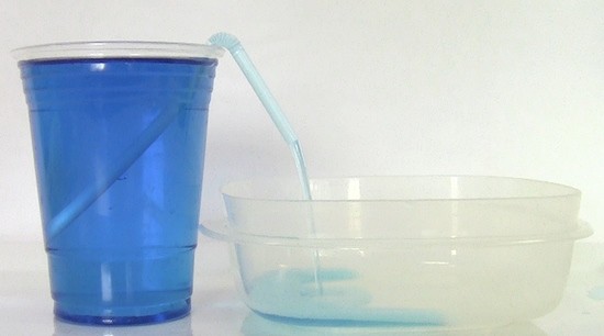 A drinking straw siphoning water out of a plastic cup into a shallow container. 