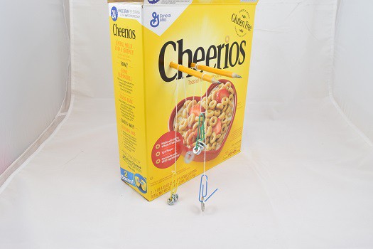 Paperclips with weights connected by string, hanging from pencils that are punctured into a cereal box