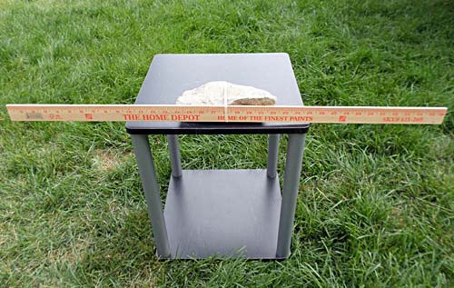 On a small table a meterstick is placed parallel to the ground and secured to a large rock