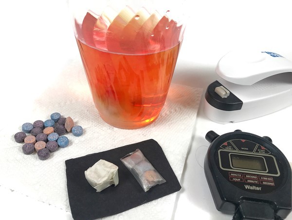 Bath tablets, built example tablets and a stopwatch next to a cup of water that has a dissolving example tablet in it.