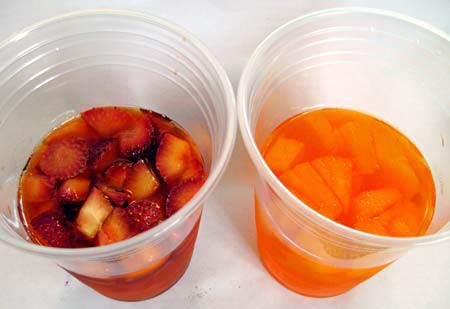 Plastic cup filled with gelatin and pieces of strawberry next to another cup filled with gelatin and pieces of pineapple