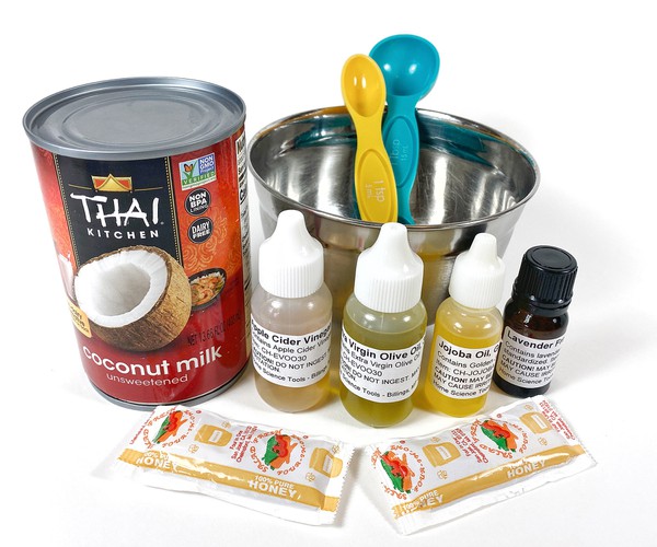 A can of coconut milk, several small bottles filled with liquids, 2 packets of honey, and a bowl with two measuring spoons on a table. 