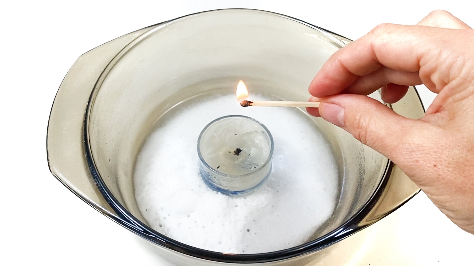 the image shows a candle insight a glass bowl. Around the candle, there is white foam. A hand is holding a lit match above the candle.