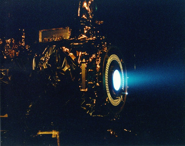 An ion engine with a jet of glowing blue atoms being emitted 