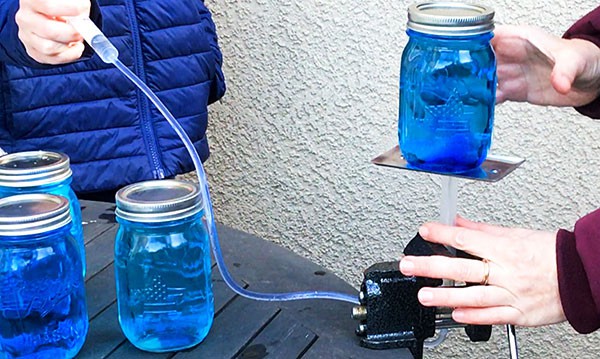 Person pushing the primary syringe of a hydraulic lift in. A jar filled with water is placed on the platform of the hydraulic lift. A person holds the second syringe upright and is ready to catch the mason jar in case it would tumble. 