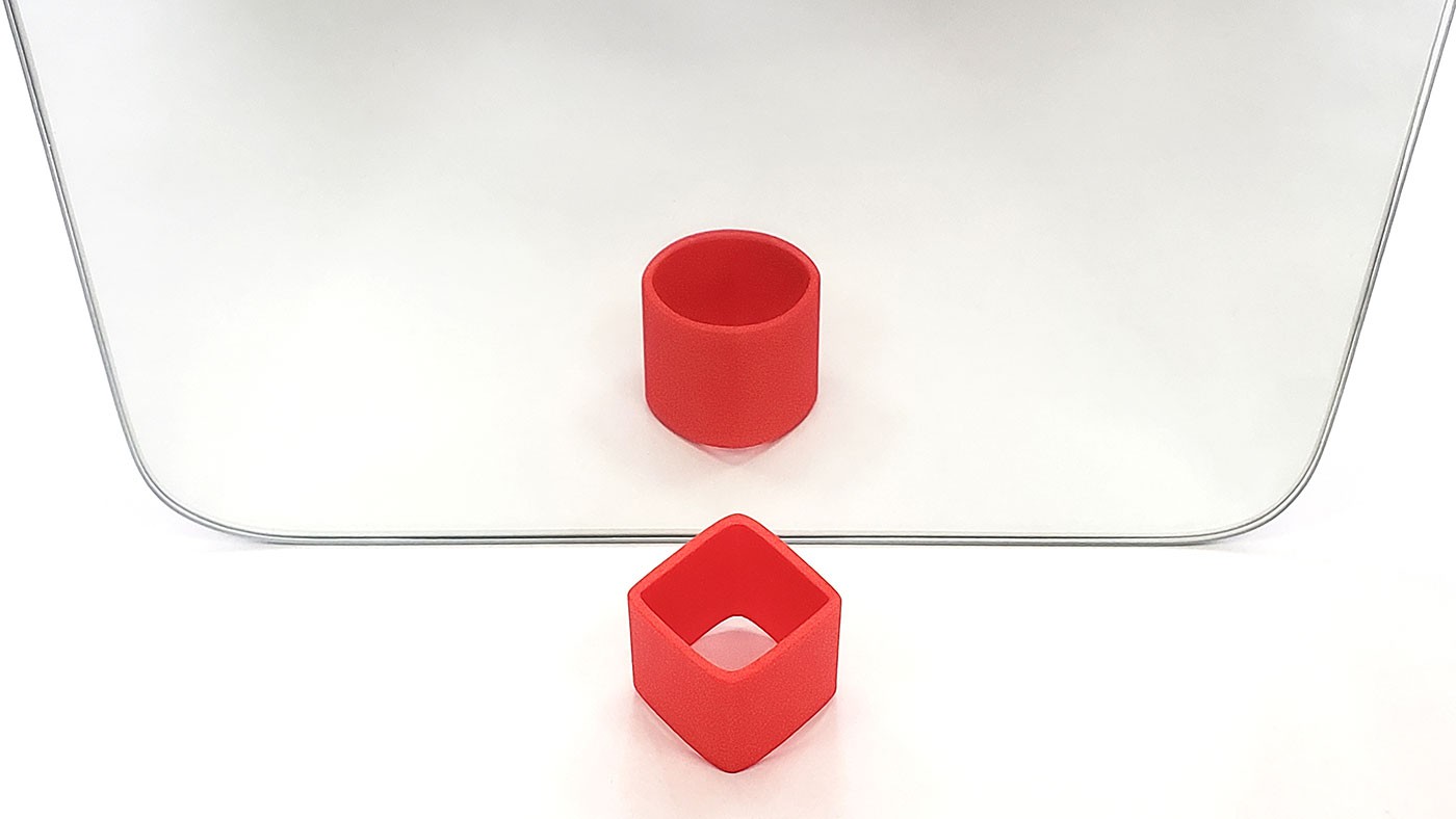 A 3D printed cylinder in front of a mirror. The cylinder looks like it has a square shape, but its reflection in the mirror looks like a circle.