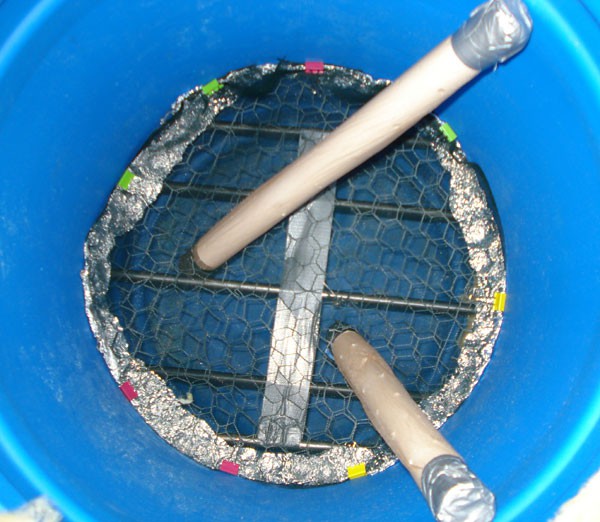 A chicken wire and mesh circle lie on top of metal rods at the bottom of a plastic barrel