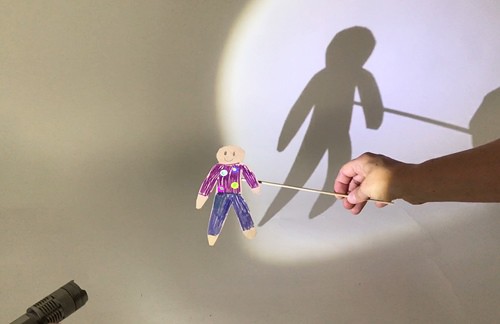 Hands holding a flashlight and a cardstock puppet to cast a shadow of the puppet. The flashlight shines onto the puppet from the side.