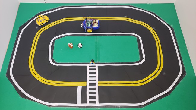 A two-lane, oval track made of black paper with lane lines and a crosswalk. Toys represent obstacles. 