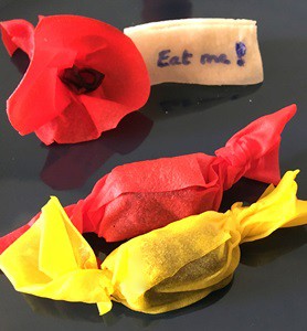 A flour from red edible paper, a name tag with the words 'Eat me'  and two candies wrapped in red and yellow edible paper.  