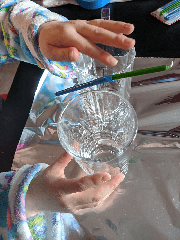 Two hands carefully balancing candles that have been taped end to end over the gap between two same-sized drinking glasses