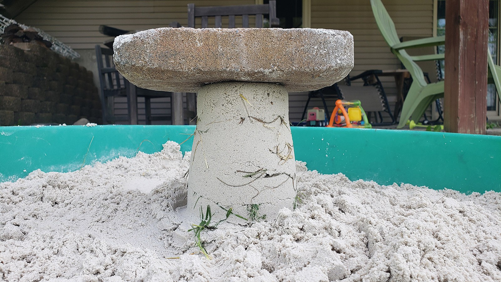 A bucket-shaped sandcastle in a sandbox with some pieces of grass sticking out the sides and a brick resting on top. 