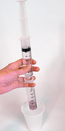 A syringe filled with alcohol is held over a syringe filled with pink sand over a plastic cup