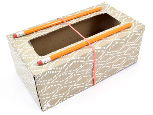 A rubber band holds two pencils in parallel next to the opening of an empty tissue box