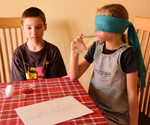 A child sits at a table with another child who is blindfolded and is taking a sip of water