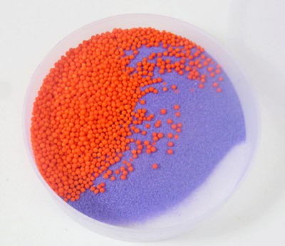 A layer of small orange beads sit above a layer of purple sand in a petri dish