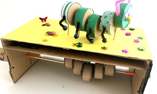 A side view of a caterpillar cardboard automaton. The body of the caterpillar is made up of 5 short cylinders, each one attached to a skewer that sticks out of a box. Inside the box, we see five cylinders attached to an axle. Each cylinder in the box is positioned under one caterpillar body part.  