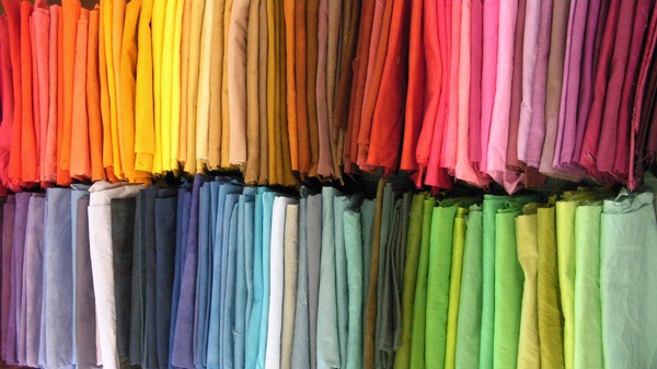 Two rows of dozens of fabrics dyed in many solid shades of red, orange, yellow, brown, blue, green, and purple.