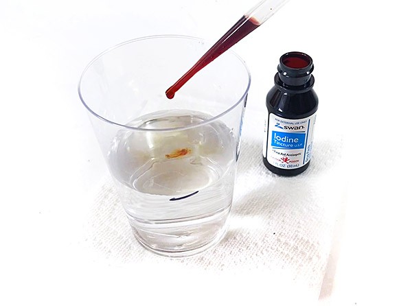 A drop of iodine solution is falling into a cup of water from a pipette tip.