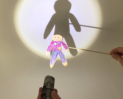 Hands holding a flashlight and a cardstock puppet to cast a shadow of the puppet.