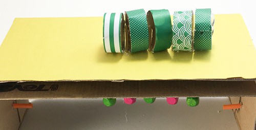 A cardboard box with five short cylinders glued to sticks sitting on top.  The box has a second cardboard panel. 5 beads are visible below the second panel, one under each body part. 