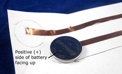 A coin cell battery is placed on a strip of copper tape on a piece of paper