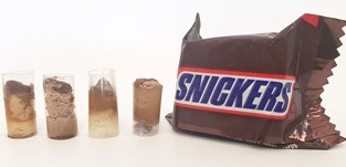 Four 'core samples' of candy bars inside short sections of clear drinking straw, next to a mini Snickers bar.