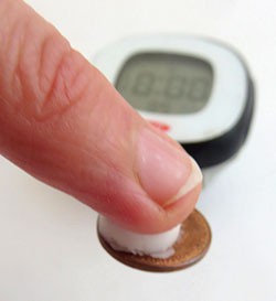 A small piece of lip balm is pressed between a fingertip and a penny