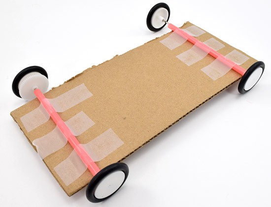 Two axles are taped to a rectangular piece of cardboard which will serve as the chassis for a solar powered car