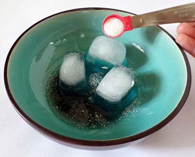A measuring spoon is used to evenly sprinkle salt over the three ice cubes arranged in the bowl