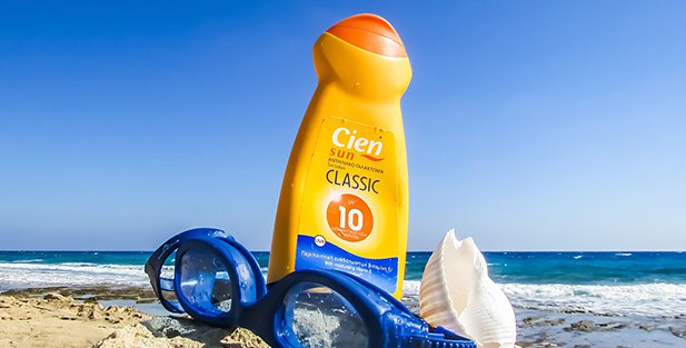  Sunscreen lotion and swimming goggles on the beach on front of the ocean.