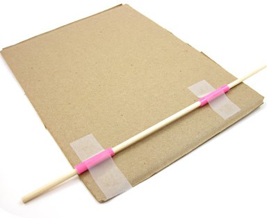 A wooden skewer inserted through two pieces of plastic straws is taped to a sheet of cardboard