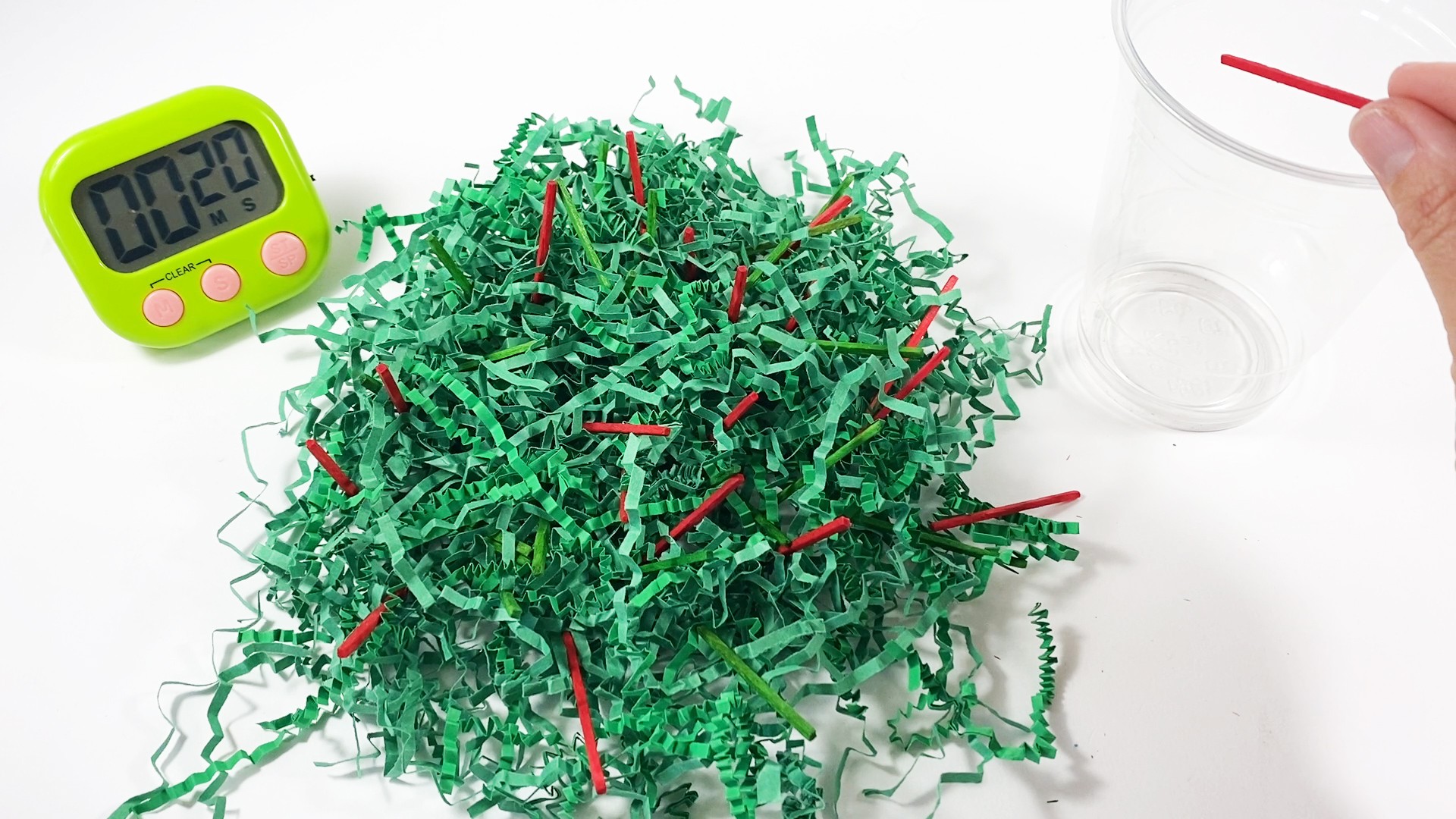 Green paper shreds with green and red matchsticks inside. A timer shows 20 seconds. A hand places a red stick into a cup.