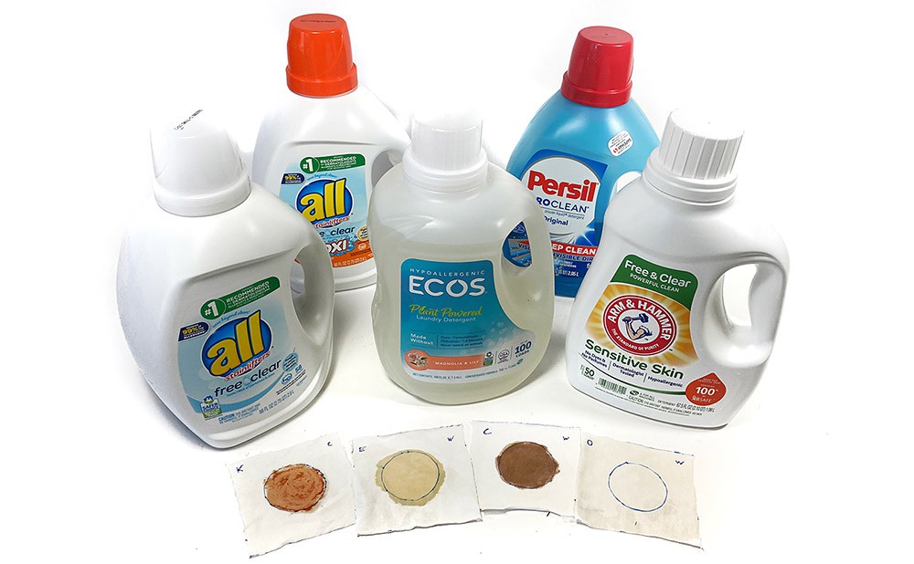 Five different laundry detergent bottles and four stained squares of cotton fabric.