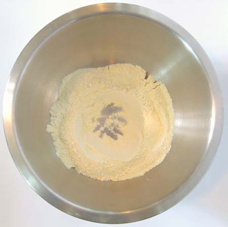 An indent is made at the center of a pile of dry baking ingredients in a metal bowl