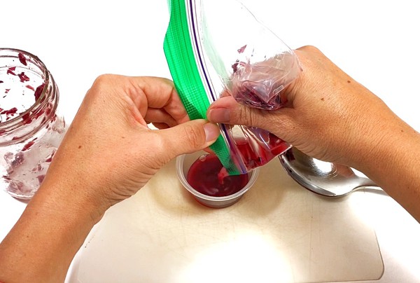 A person is holding a small resealable bag on top of a small cup. Inside the bag is a mixture of cut red leaves and isopropyl alcohol. The red leaf extract is pouring from an opening of the bag into the small cup.