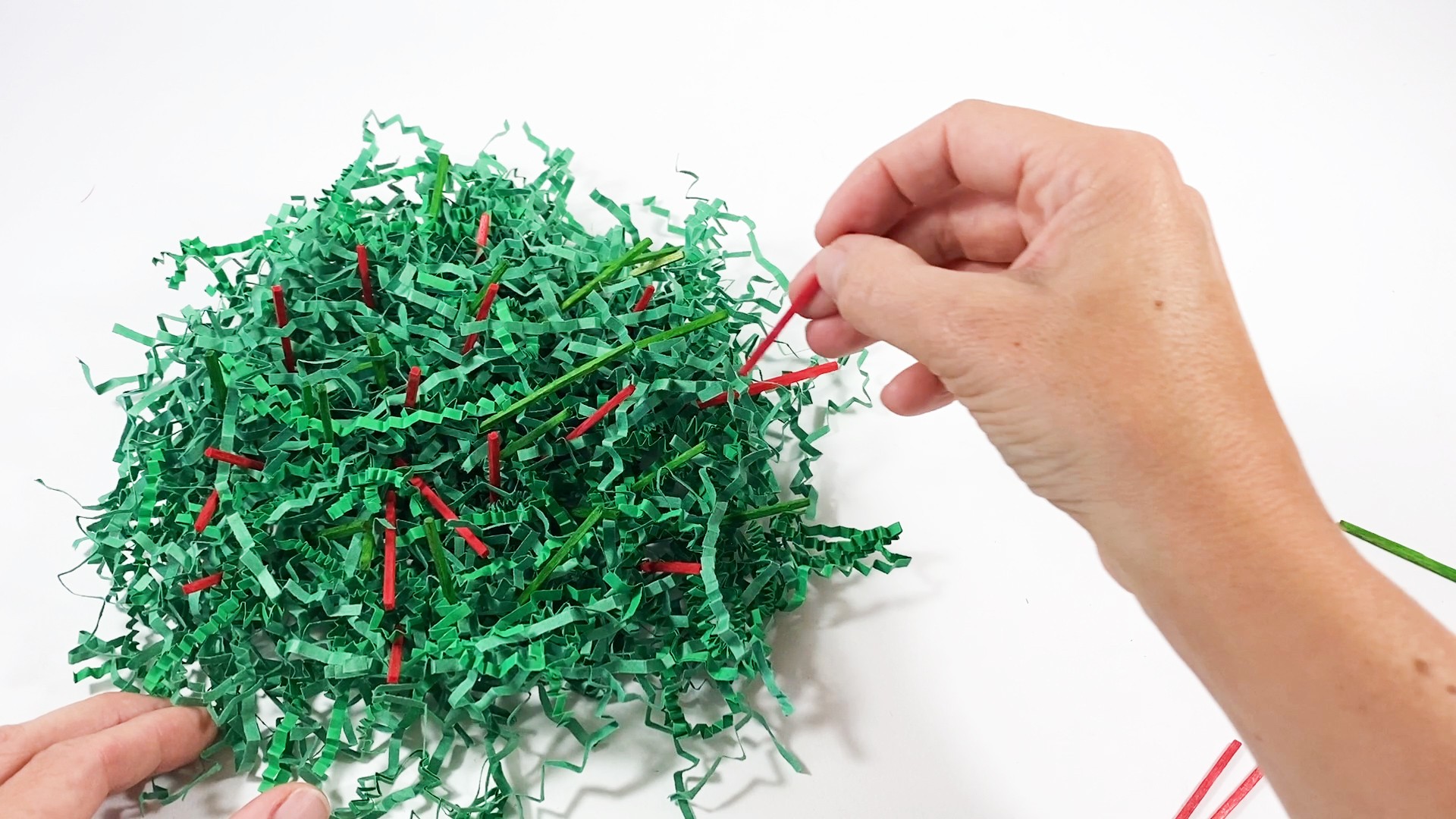 Green paper shreds with green and red matchsticks hidden inside. A hand places a red matchstick into the paper shreds.
