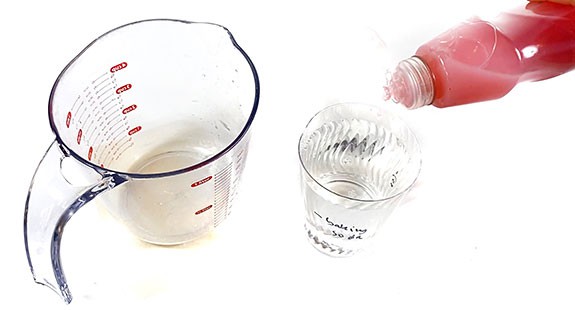 A measuring cup is standing next to a cup filled with water. One drop of dish soap is added to the cup.
